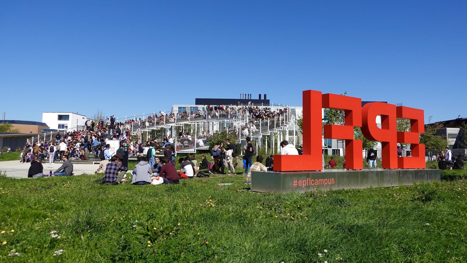 Vue sur l'Agora Lombard Odier, à l'EPFL. Des membres de l'EPFL profitent du soleil pour manger à l'extérieur sur les marches. A droite de l'image, un totem avec le logo de l'EPFL - 2022 EPFL/Xurxo Adrián Entenza - CC-BY-SA 4.0