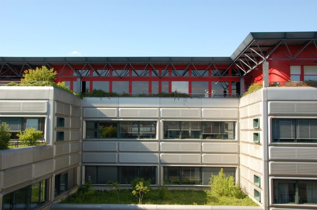 Détail d'un bâtiment de la Faculté de Génie Civil couvert en façade de panneaux métalliques gris qui alternent avec des fenêtres allongées et qui possèdent des avant-corps aux toits végétalisés. La toiture du bâtiment est faite d'une structure métallique bleue.