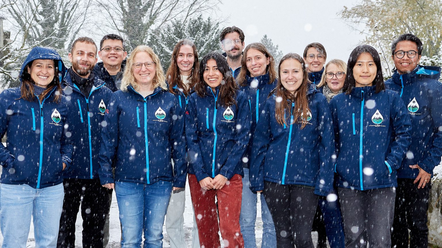 13 members of LEV team standing in dark blue LEV jackets outdoors as the snow falls