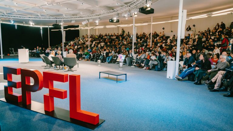 Le logo EPFL posé dans le Forum Learning Center. En arrière-plan, un public assis venu assister à une conférence de la Journée Intelligence Artificielle, le 23 novembre 2024 © EPFL / Adrien Buttier